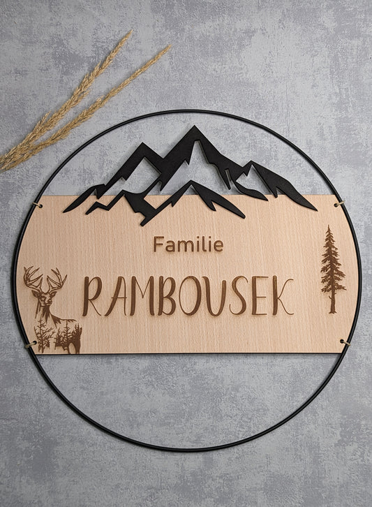 Personalisierte Wanddeko mit Holzschild, graviertem Familiennamen 'Rambousek', Bergsilhouette und Naturmotiven wie Hirsch und Baum. Eingefasst in einen schwarzen Metallring, präsentiert auf grauem Hintergrund mit Trockenpflanzen.
