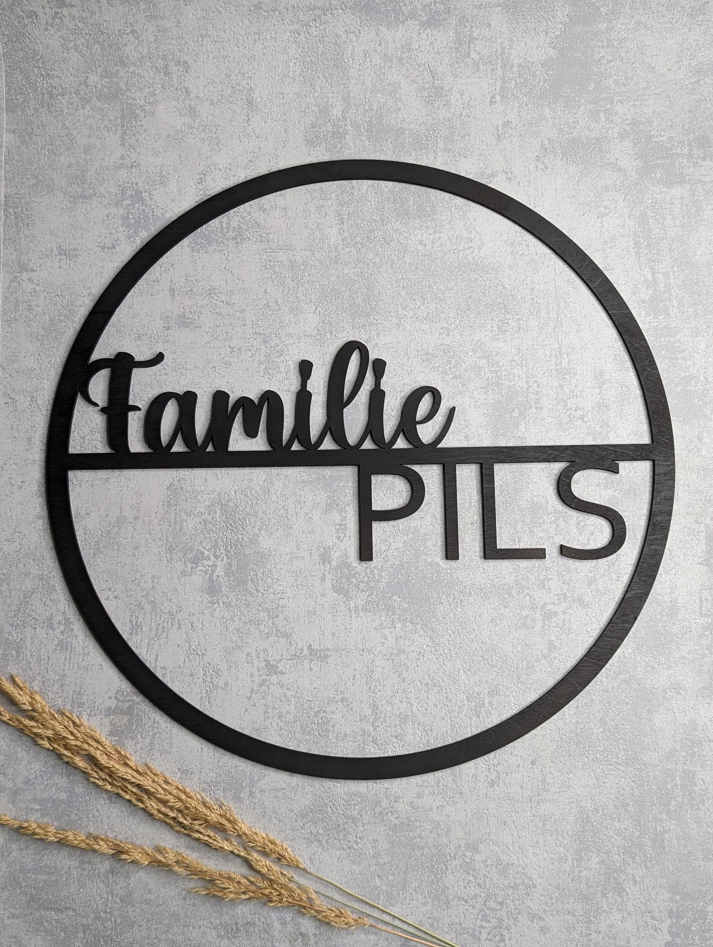 Schwarzes Holzschild im Kreisdesign mit dem Schriftzug 'Familie Pils'. Moderne Wanddekoration, präsentiert auf grauem Hintergrund mit Trockenpflanzen.