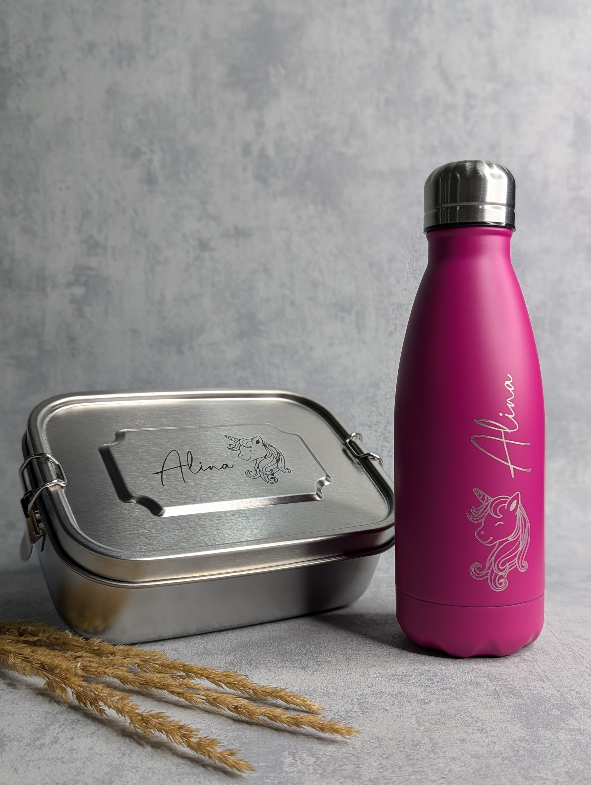 Personalisierte Edelstahl-Lunchbox mit Namensgravur 'Alina' und Einhorn-Motiv, passend dazu eine pinke Thermo-Trinkflasche mit gleicher Gravur. Beide Artikel stehen auf einer hellgrauen Oberfläche mit Trockenpflanzen als Dekoration.