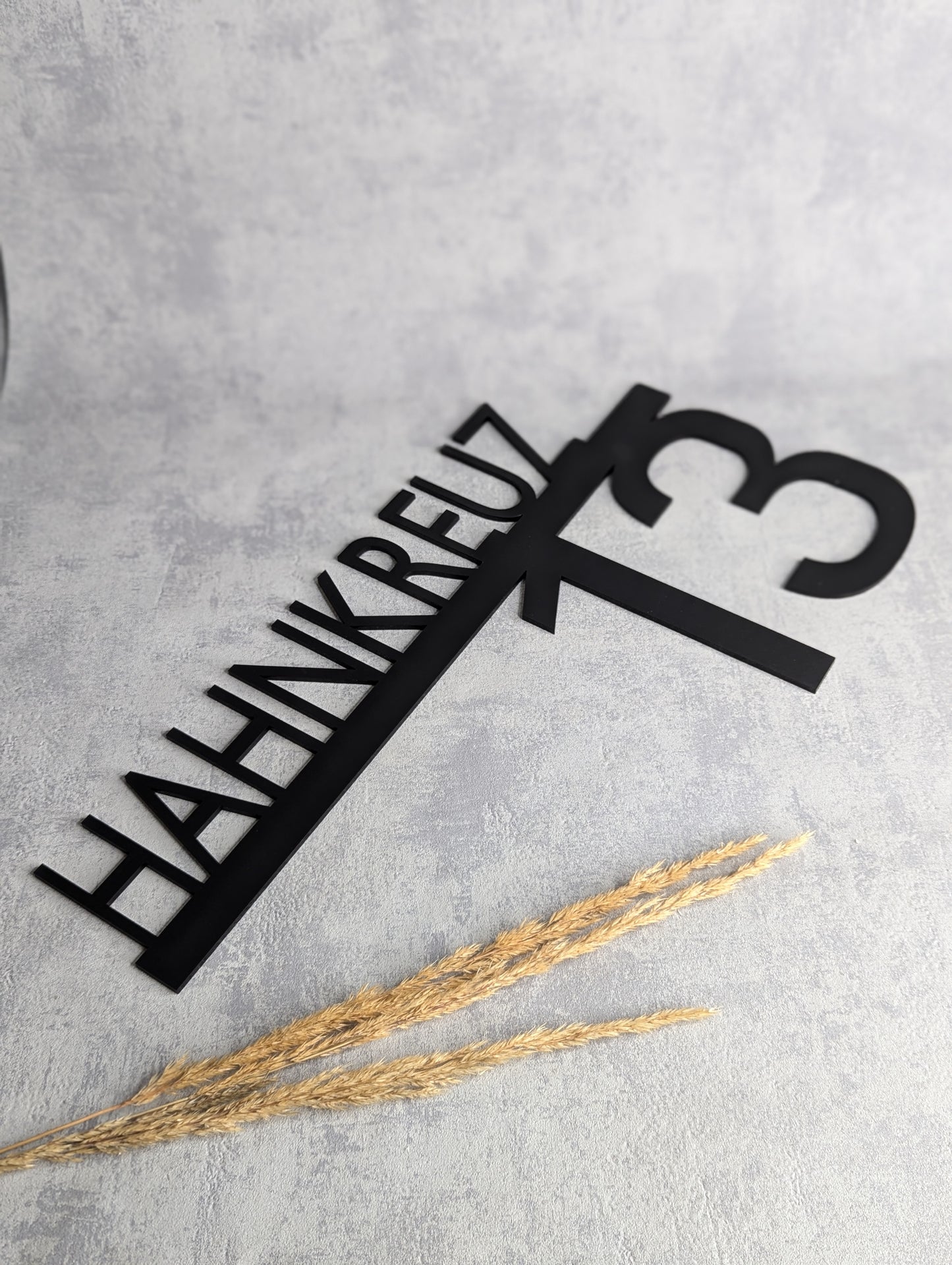 "Schwarzes, individuell gefertigtes Hausschild mit dem Schriftzug 'HAHNKREUZ' und der Hausnummer '13' in modernem Design. Das Schild liegt auf einer hellgrauen Oberfläche, mit Trockenpflanzen als Dekoration im Hintergrund."