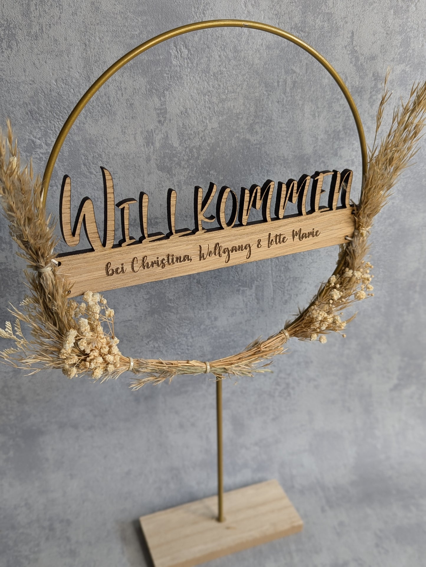 Dekorativer Willkommensring mit personalisierter Holzgravur 'Willkommen bei Christina, Wolfgang & Lotte Marie'. Der goldene Metallring ist mit Trockenblumen verziert und steht auf einem Holzsockel. Hintergrund in hellem Grau.