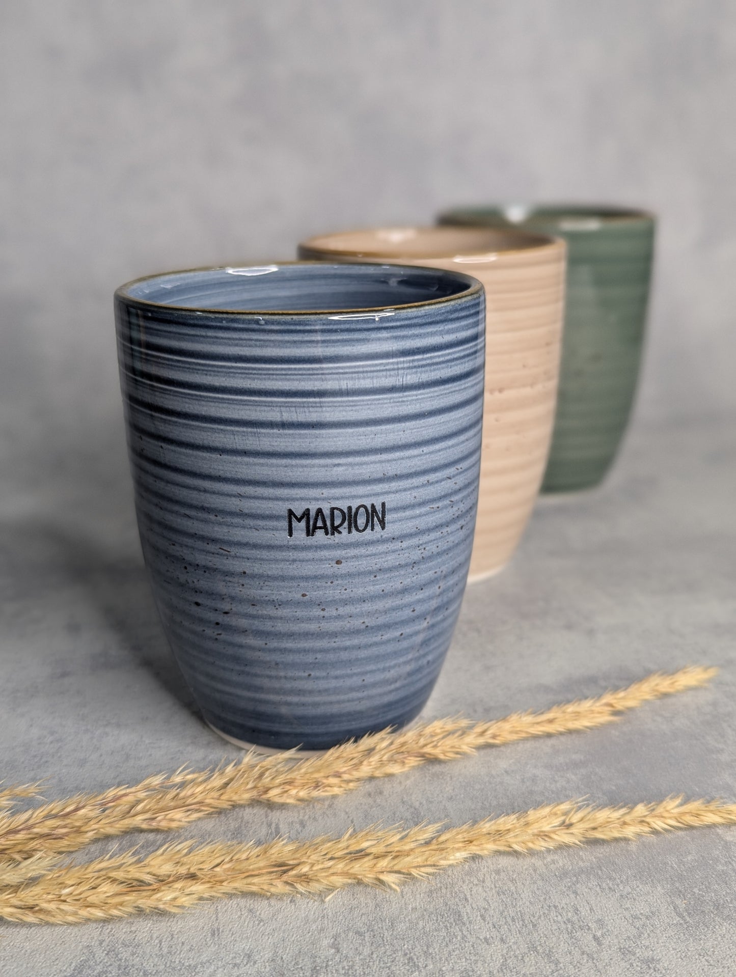 Personalisierter Keramikbecher mit Namensgravur, blau, mit zwei weiteren Bechern in beige und grün im Hintergrund.