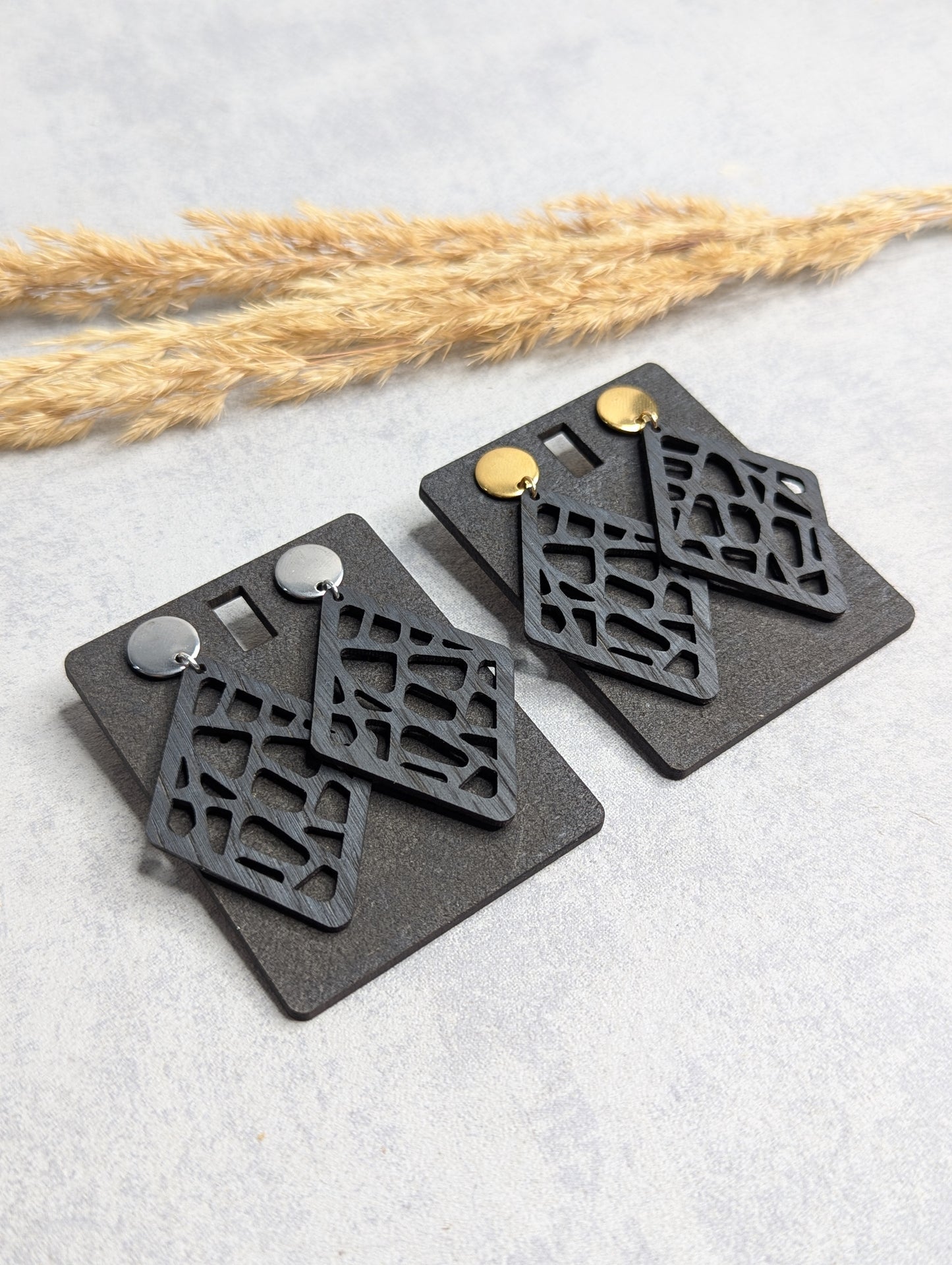 Zwei Paar geometrische Ohrringe mit filigranem, rautenförmigem Design aus schwarzem Material, befestigt an schwarzen Karten mit je einem goldenen und silbernen Steckverschluss. Hintergrund mit natürlicher Dekoration aus Trockenpflanzen auf einer hellgrauen Oberfläche."