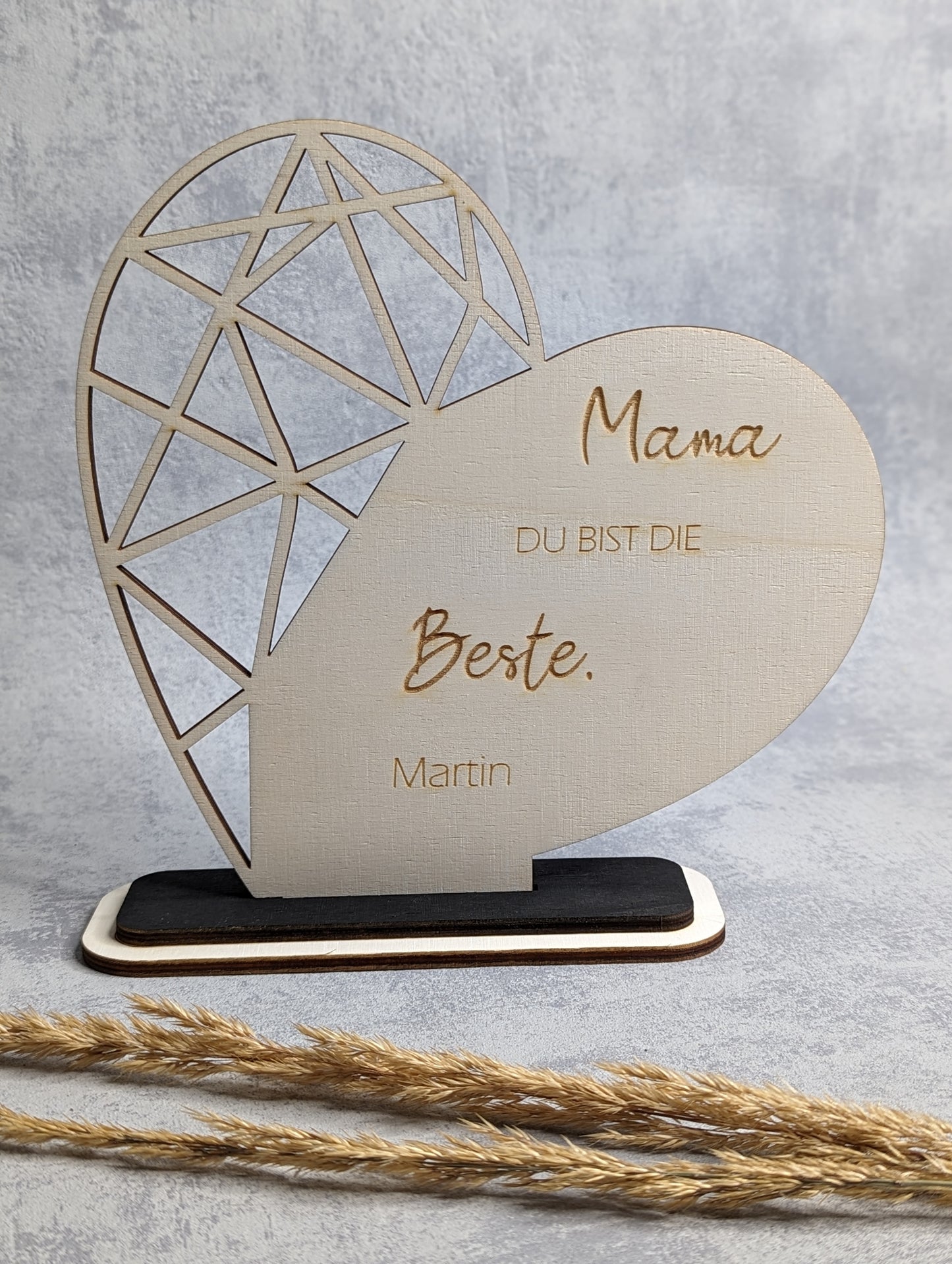 Personalisiertes Herz aus Holz mit Gravur 'Mama, du bist die Beste' und dem Namen 'Martin'. Das Herz hat ein filigranes geometrisches Design und steht auf einem dunklen Holzsockel. Präsentiert auf grauem Hintergrund mit Trockenpflanzen.