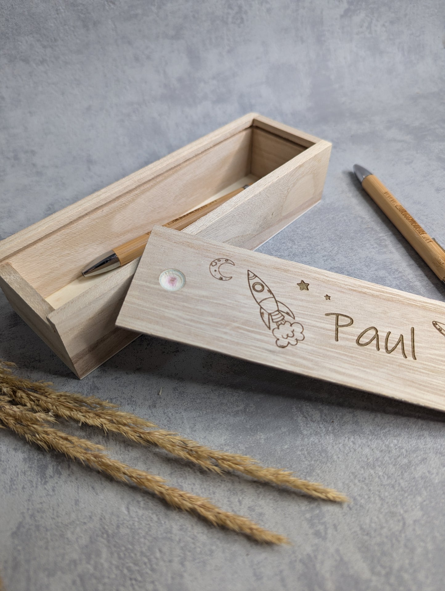 Personalisierte Holz-Stiftebox mit Schiebedeckel, graviert mit dem Namen 'Paul' und einer Rakete mit Sternen und Mond. Die Box ist geöffnet und enthält einen Bambus-Kugelschreiber. Eine zweite Stiftebox liegt daneben auf einer hellgrauen Oberfläche mit Trockenpflanzen als Dekoration.