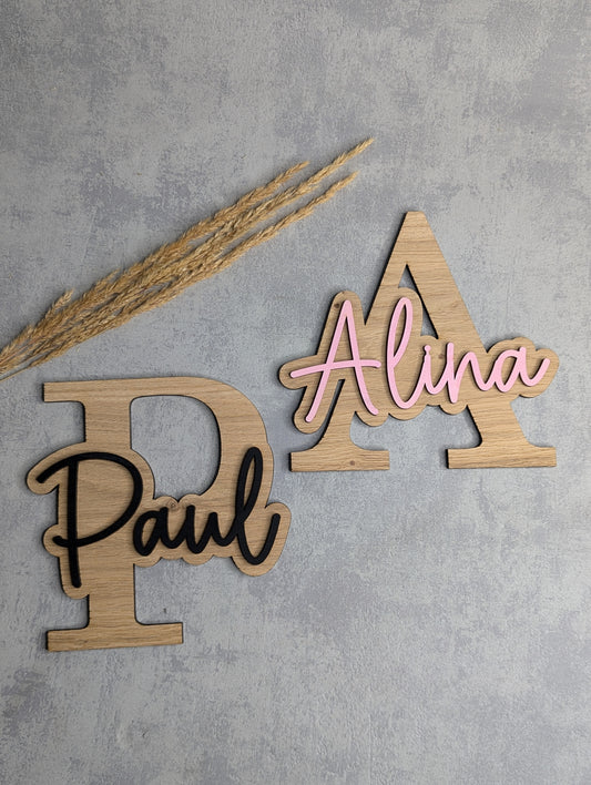 Zwei personalisierte Holz-Namensschilder mit großen Buchstaben 'P' und 'A'. Die Namen 'Paul' in Schwarz und 'Alina' in Rosa sind darauf in einer verspielten Schrift befestigt. Die Schilder liegen auf einer hellgrauen Oberfläche mit Trockenpflanzen als Dekoration.