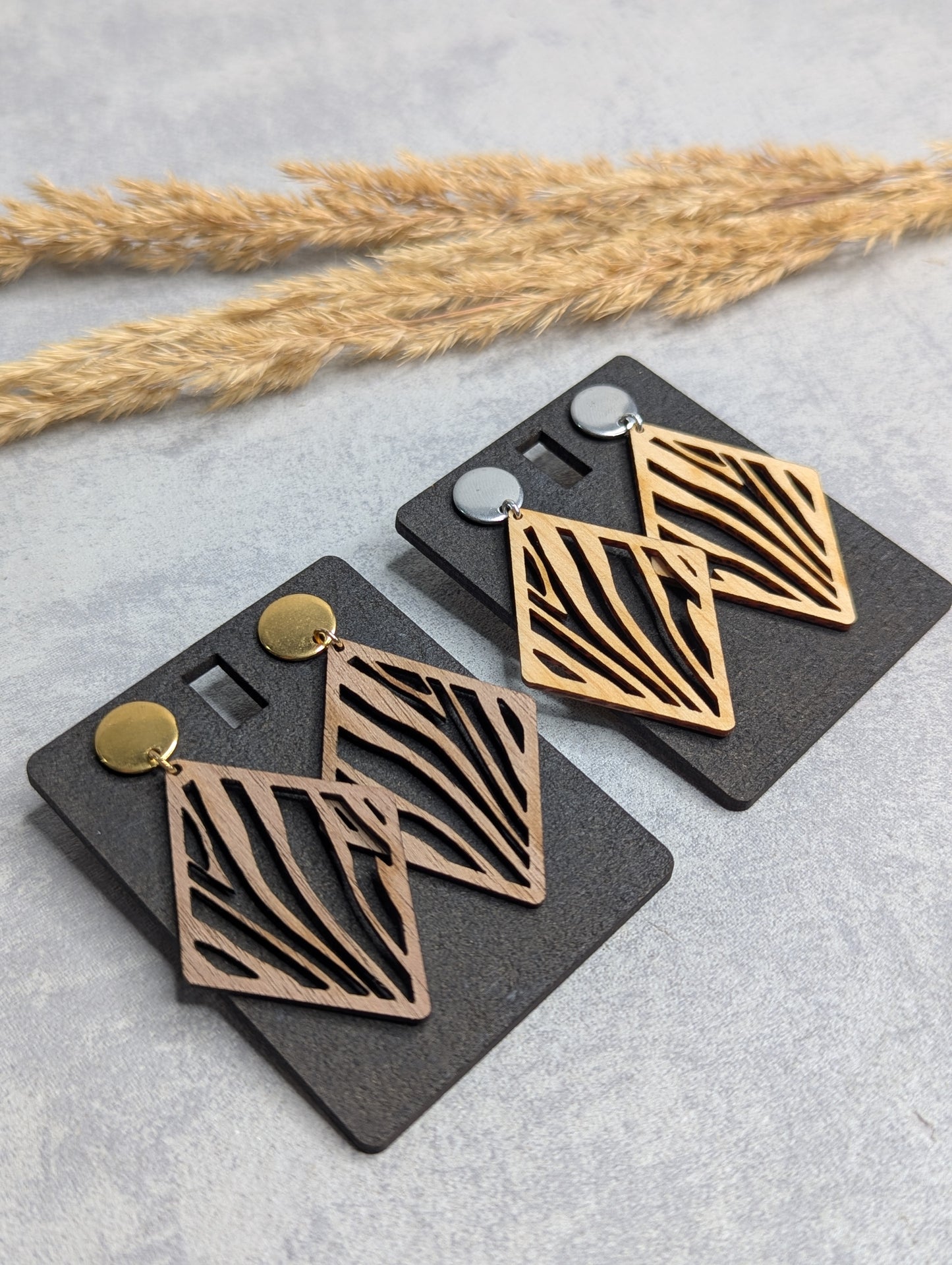 "Zwei Paar hölzerne Ohrringe mit rautenförmigem Design und filigranem Zebra-Muster, befestigt an schwarzen Karten mit je einem goldenen und silbernen Steckverschluss. Hintergrund mit Trockenpflanzen auf einer hellgrauen Oberfläche."