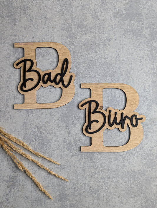 Zwei dekorative Türschilder aus Holz mit großen Buchstaben 'B' und aufgesetzten Schriftzügen 'Bad' und 'Büro' in einer stilvollen Kombination aus Holz und schwarzer Schrift. Die Schilder liegen auf einer hellgrauen Oberfläche, mit Trockenpflanzen als Dekoration.