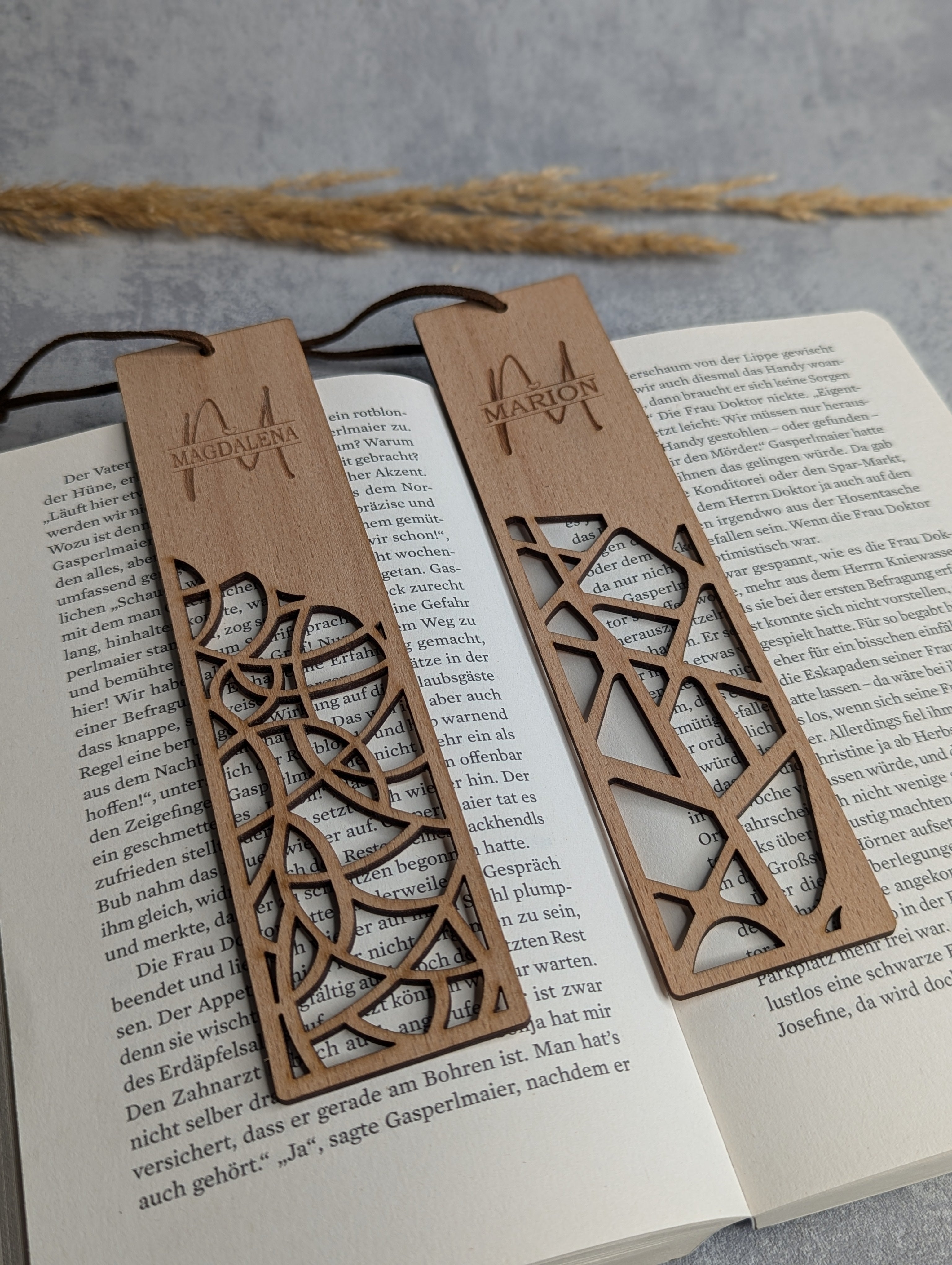 Zwei personalisierte Holz-Lesezeichen mit filigranen Mustern und den eingravierten Namen 'Magdalena' und 'Marion'. Die Lesezeichen liegen auf einem geöffneten Buch und sind mit einer braunen Kordel versehen.