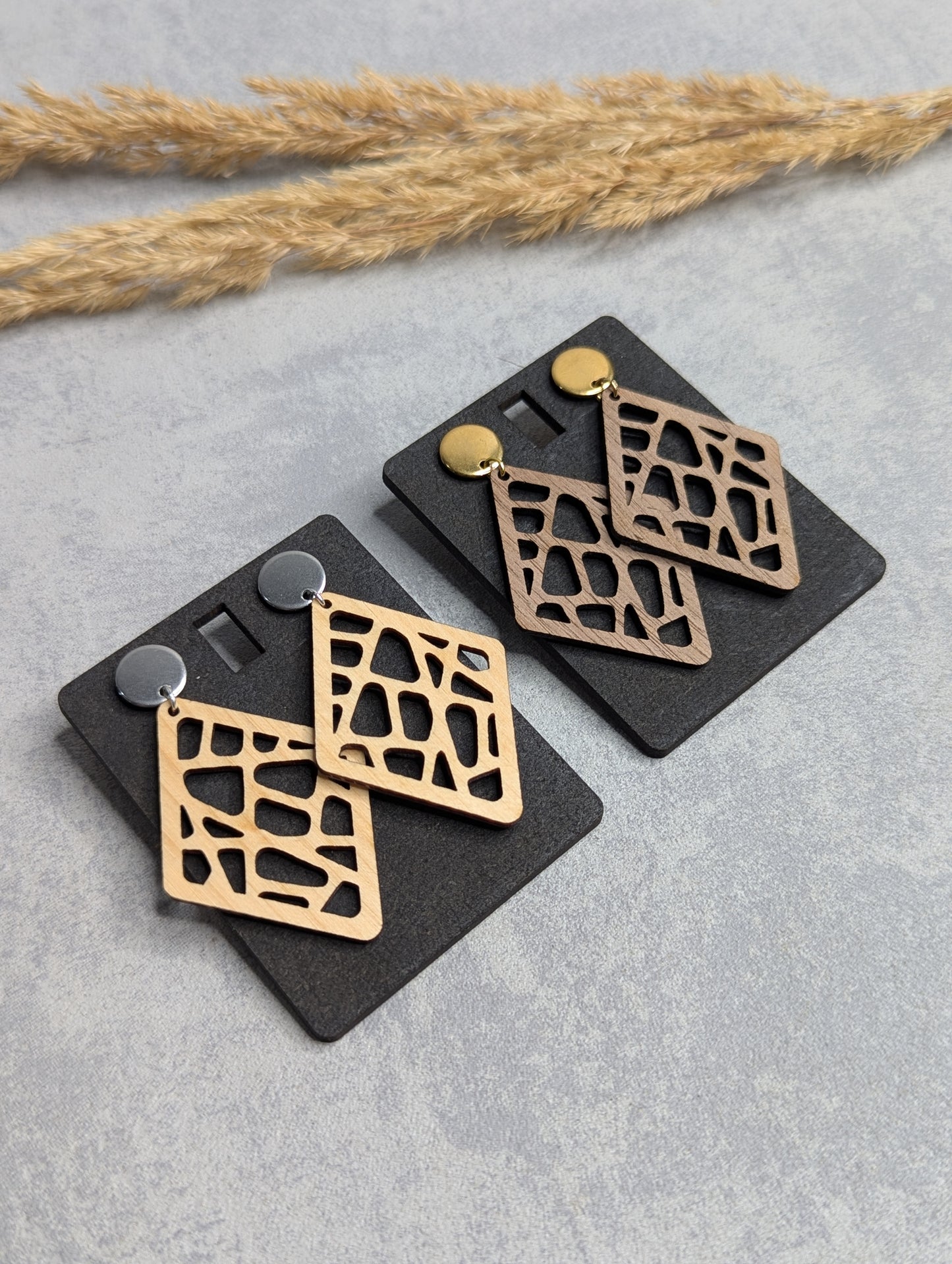 "Zwei Paar hölzerne Ohrringe mit rautenförmigem Design und einem filigranen Muster, das an eine Giraffenfell-Struktur erinnert. Die Ohrringe sind an schwarzen Karten befestigt, mit je einem goldenen und silbernen Steckverschluss. Der Hintergrund zeigt eine hellgraue Oberfläche mit Trockenpflanzen als Dekoration."