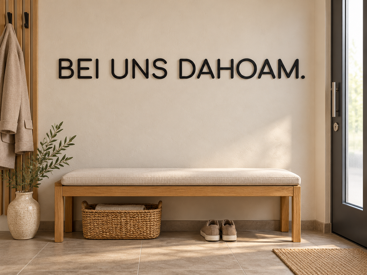 BEI UNS DAHOAM. als einzeiliger schwarzer 3D-Schriftzug in Comfortaa über einer Holzbank im gemütlichen Hausvorraum