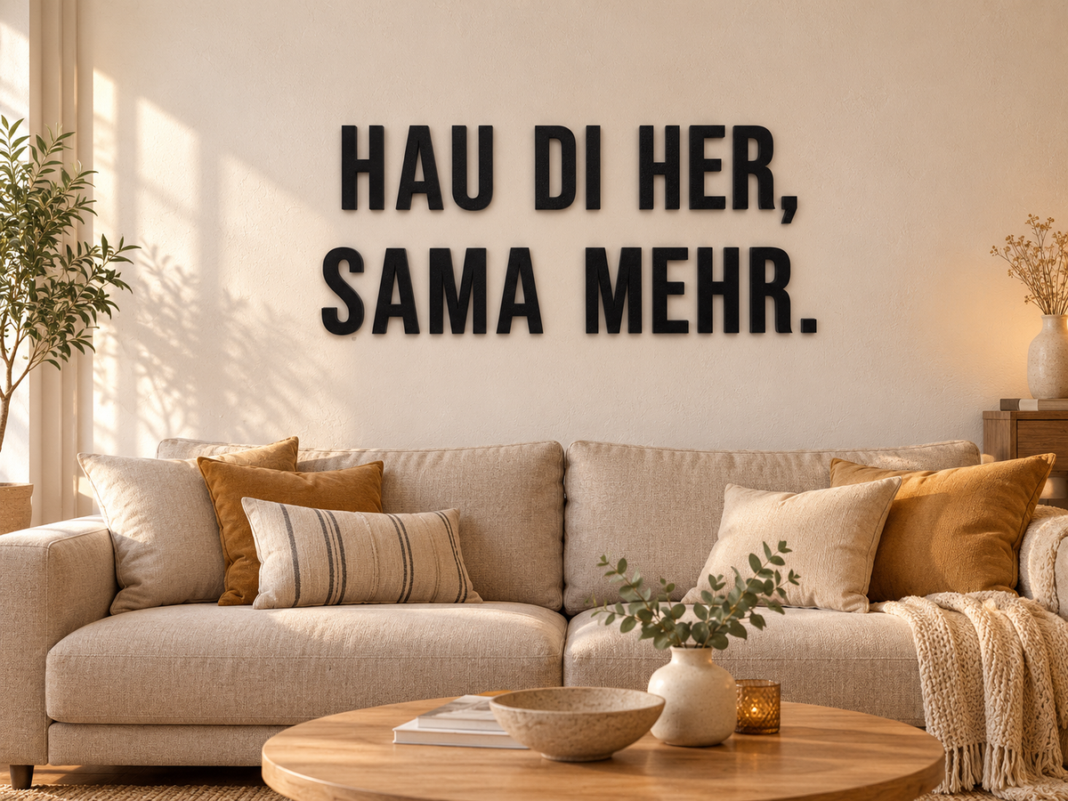 Österreichischer Dialektspruch „HAU DI HER, SAMA MEHR.“ aus schwarzen 3D-Buchstaben über einem beigen Sofa