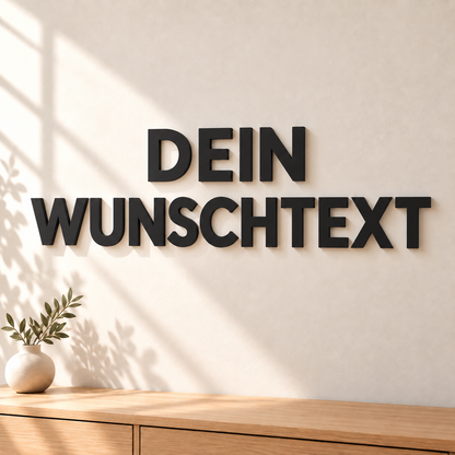 Personalisierte schwarze 3D-Wandbuchstaben „DEIN WUNSCHTEXT“ an einer hellen Wand über einer Holzkommode