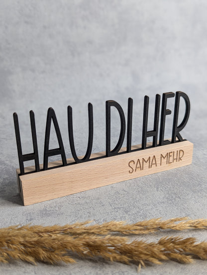 Mundart-Holzaufsteller mit dem Spruch 'HAU DI HER' in schwarzen Holzbuchstaben und der Gravur 'SAMA MEHR' im Holzsockel. Rustikale, handgefertigte Deko aus Naturholz, präsentiert auf grauem Hintergrund mit Trockenpflanzen als Dekoration.