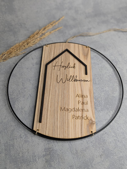 Personalisiertes Holzschild mit Willkommen-Schriftzug und Familiennamen im Metallring auf grauem Hintergrund