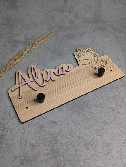 "Personalisierte Holzgarderobe mit dem Namen 'Alina' in rosa Schrift und einer eingravierten Einhorn-Grafik. Die Garderobe hat zwei schwarze Haken und besteht aus hellem Holz. Sie liegt auf einer hellgrauen Oberfläche mit Trockenpflanzen als Dekoration im Hintergrund."