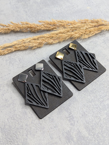 Zwei Paar geometrische Ohrringe aus schwarzem Holz mit filigranem Linienmuster. Präsentiert auf schwarzen Karten, mit Edelstahl-Steckern in Silber und Gold. Dekoriert auf hellem Hintergrund mit Trockenpflanzen.
