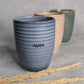 Personalisierter Keramikbecher mit Namensgravur, blau, mit zwei weiteren Bechern in beige und grün im Hintergrund.