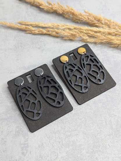 "Zwei Paar schwarze, ovale Ohrringe mit filigranem Muster, das an eine Schildkrötenpanzer-Struktur erinnert. Die Ohrringe sind an schwarzen Karten befestigt, mit je einem goldenen und silbernen Steckverschluss. Der Hintergrund besteht aus einer hellgrauen Oberfläche mit Trockenpflanzen als Dekoration."