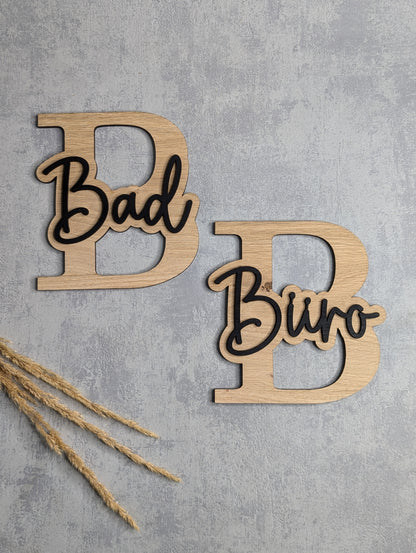 Zwei dekorative Türschilder aus Holz mit großen Buchstaben 'B' und aufgesetzten Schriftzügen 'Bad' und 'Büro' in einer stilvollen Kombination aus Holz und schwarzer Schrift. Die Schilder liegen auf einer hellgrauen Oberfläche, mit Trockenpflanzen als Dekoration.