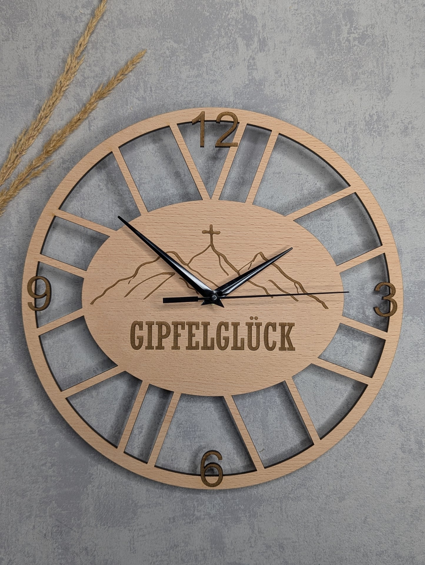 Runde Wanduhr aus hellem Holz mit dem Schriftzug 'GIPFELGLÜCK' und Bergmotiv in der Mitte, römische Ziffern 12, 3, 6, 9, schwarze Zeiger, vor grauem Betonhintergrund mit dekorativen Weizenähren