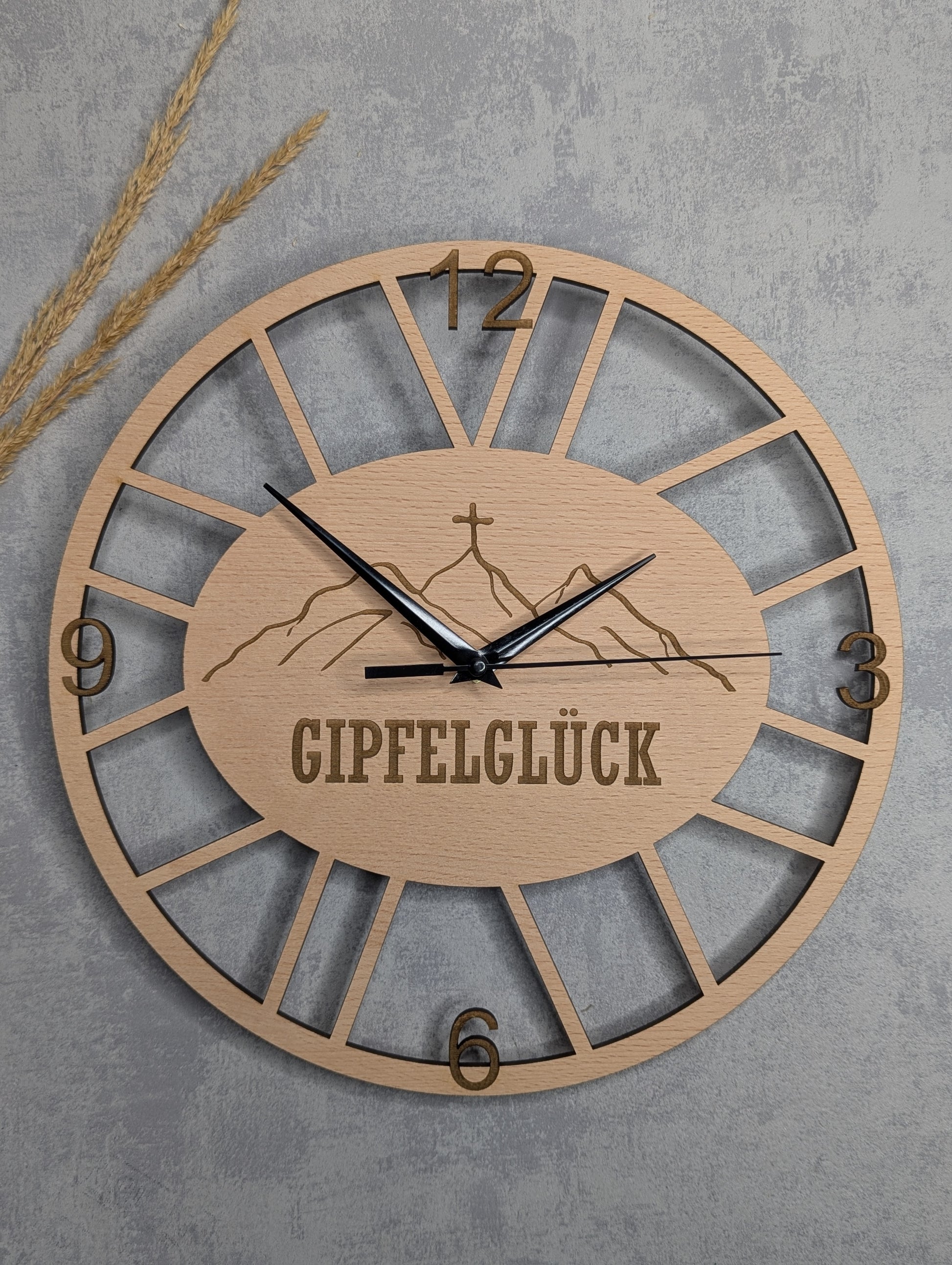 Runde Wanduhr aus hellem Holz mit dem Schriftzug 'GIPFELGLÜCK' und Bergmotiv in der Mitte, römische Ziffern 12, 3, 6, 9, schwarze Zeiger, vor grauem Betonhintergrund mit dekorativen Weizenähren