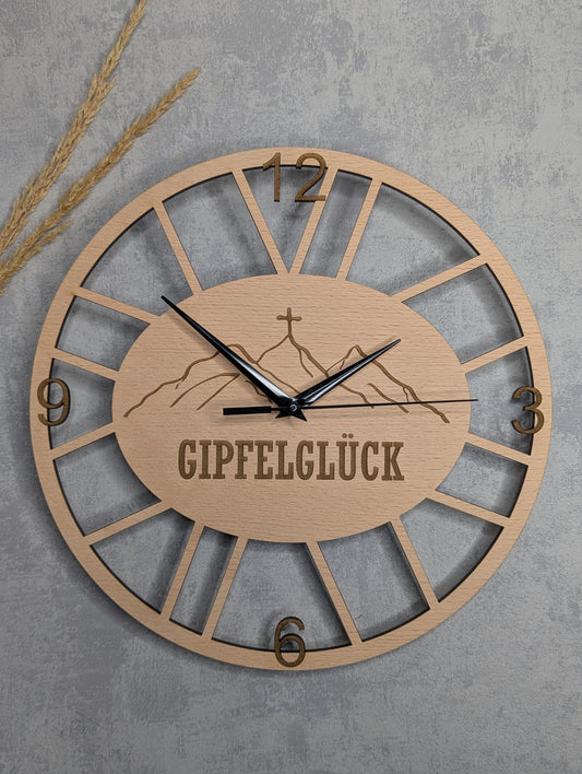 Runde Wanduhr aus hellem Holz mit dem Schriftzug 'GIPFELGLÜCK' und Bergmotiv in der Mitte, römische Ziffern 12, 3, 6, 9, schwarze Zeiger, vor grauem Betonhintergrund mit dekorativen Weizenähren